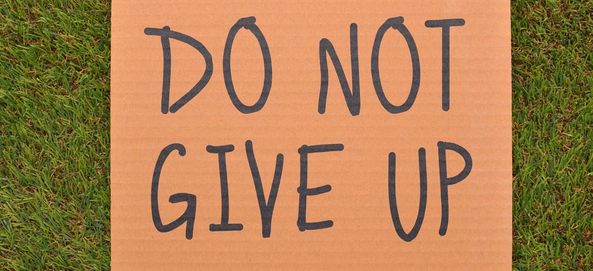 Cardboard sign reading 'Do Not Give Up' placed on green turf, symbolizing perseverance.