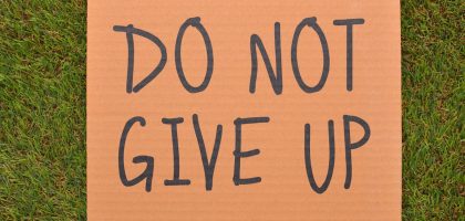 Cardboard sign reading 'Do Not Give Up' placed on green turf, symbolizing perseverance.