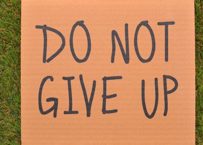 Cardboard sign reading 'Do Not Give Up' placed on green turf, symbolizing perseverance.